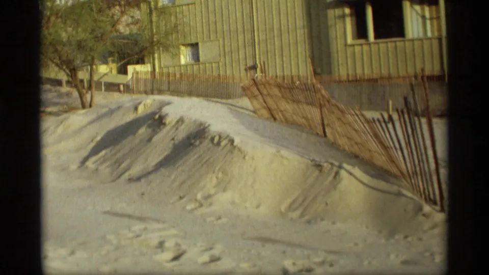 1969 Holland, Michigan beachside frame showing a sand dune, rustic fence, bare tree, and nearby sided building.