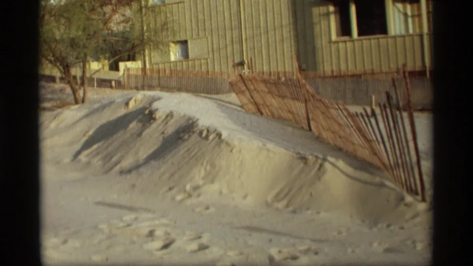 1969 Holland, Michigan home-movie view of a dune edge with footprints, leaning fencing, a bare tree, and a beachside building.