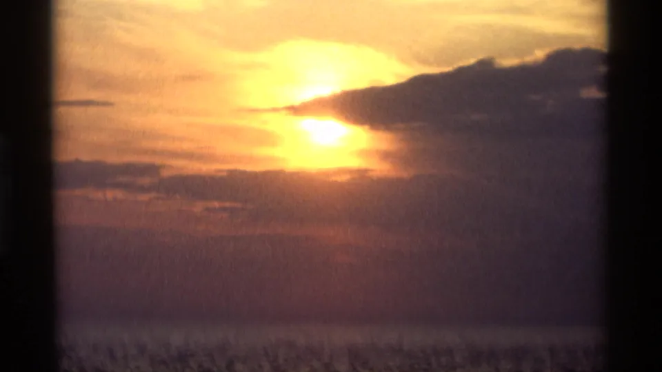 1969 Holland, Michigan sunset frame with the sun near dark clouds, warm orange light, water below, and a heavy vertical foreground bar.