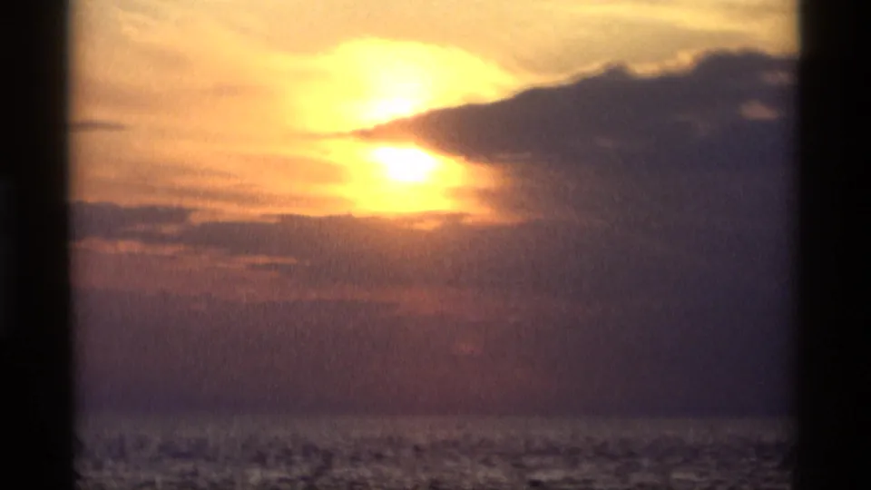 1969 Holland, Michigan home-movie frame showing a glowing sunset, layered clouds, and water below, bordered by dark vertical foreground shapes.