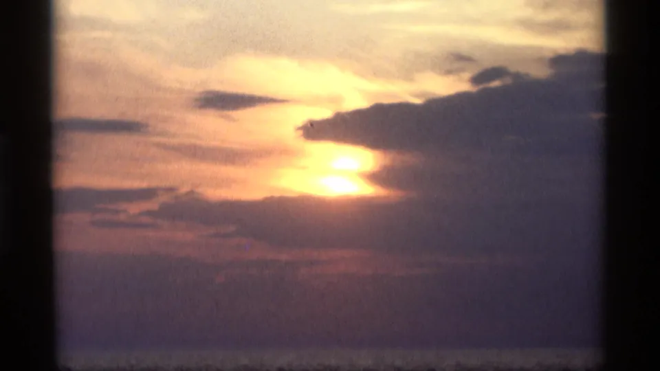 1969 Holland, Michigan frame with the sun sinking behind a dark cloud mass, warm sky color, and a faint waterline beneath.