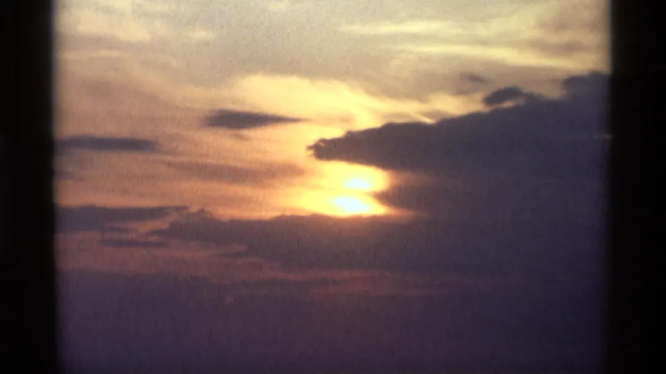 1969 Holland, Michigan sunset frame with the sun diffused through cloud cover and warm light against a blue sky.