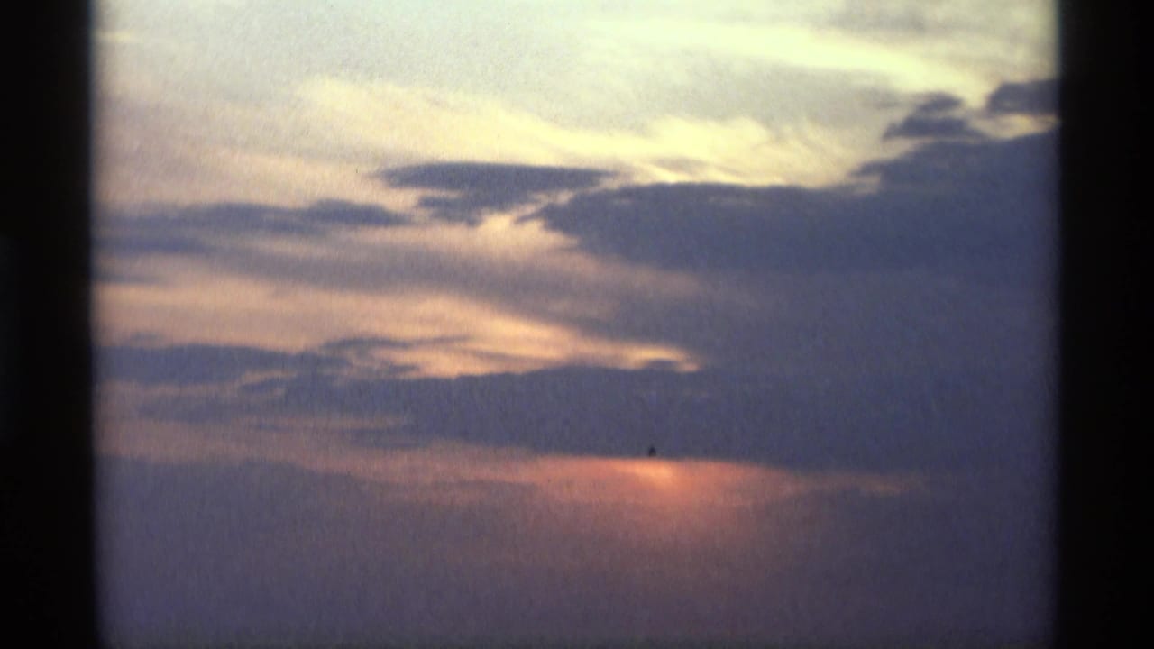 1969 8mm home-movie frame from Holland, Michigan showing a subdued sunset glow behind blue and purple clouds over a distant body of water.