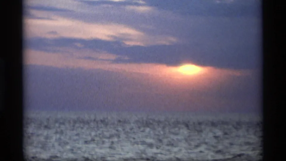 1969 8mm frame from Holland, Michigan showing the sun breaking through low clouds near the water horizon with rippled water in the foreground.