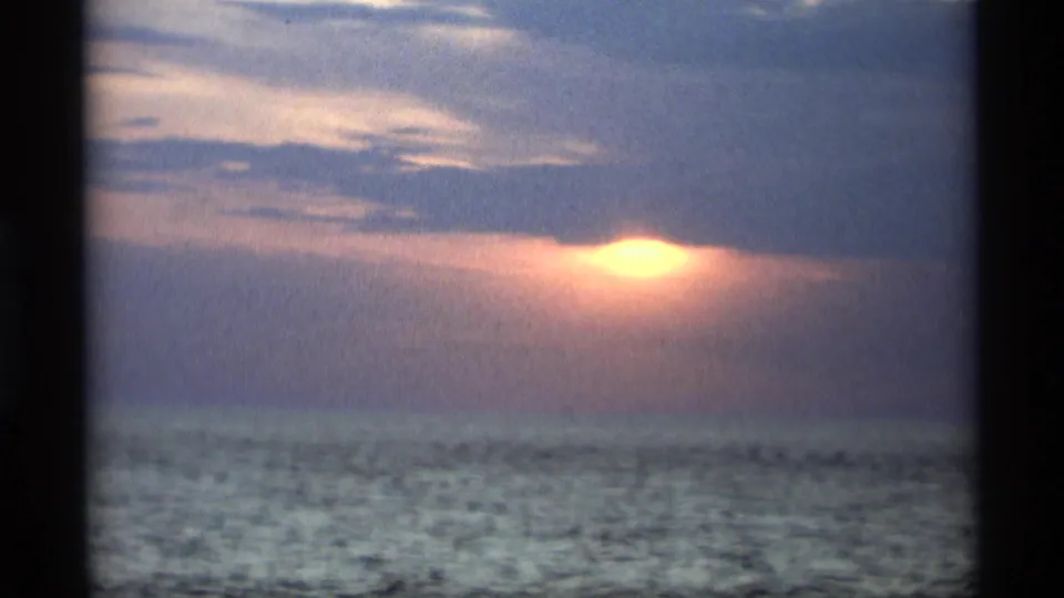 1969 Holland, Michigan home-movie still with a bright sunset opening beneath blue clouds and a darkened water surface below.