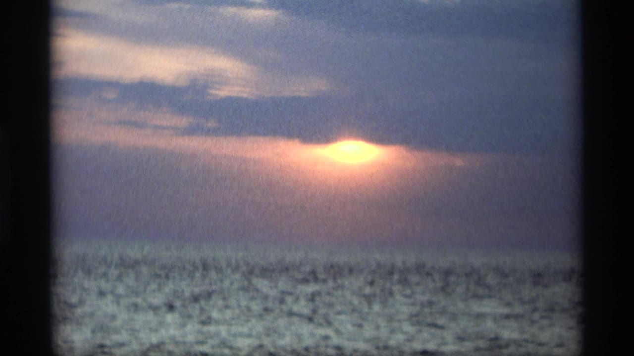 Low sun behind clouds over open water with visible film grain.