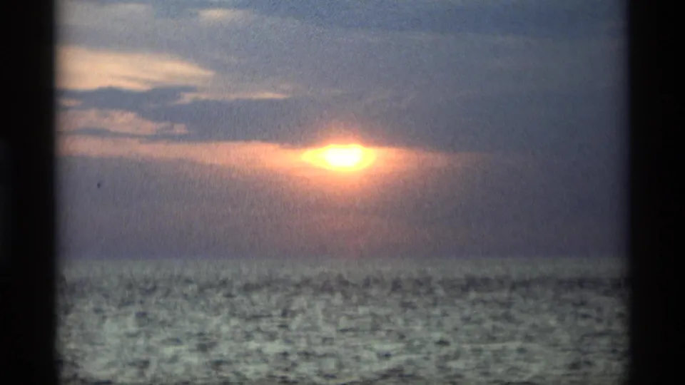 Grainy 8mm Holland, Michigan frame showing a warm sunset glow filtered by clouds above rippled open water.