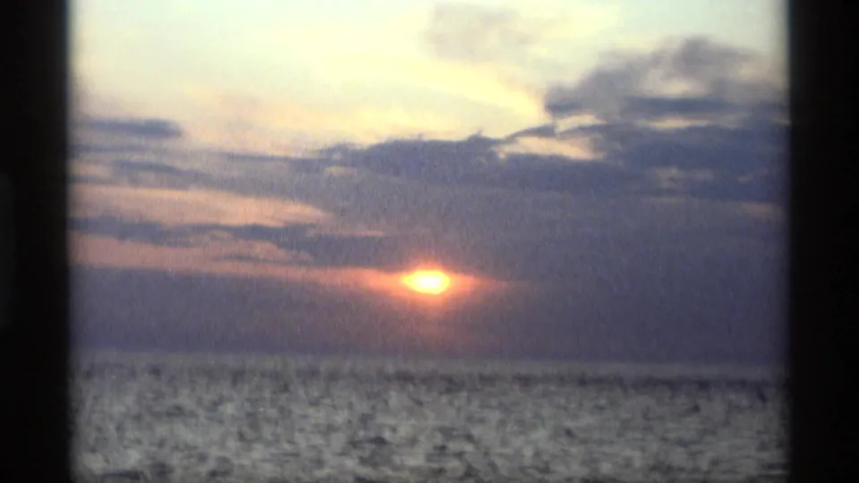 1969 home-movie frame showing a soft orange sun over water at Holland, Michigan, with clouds filling most of the sky.