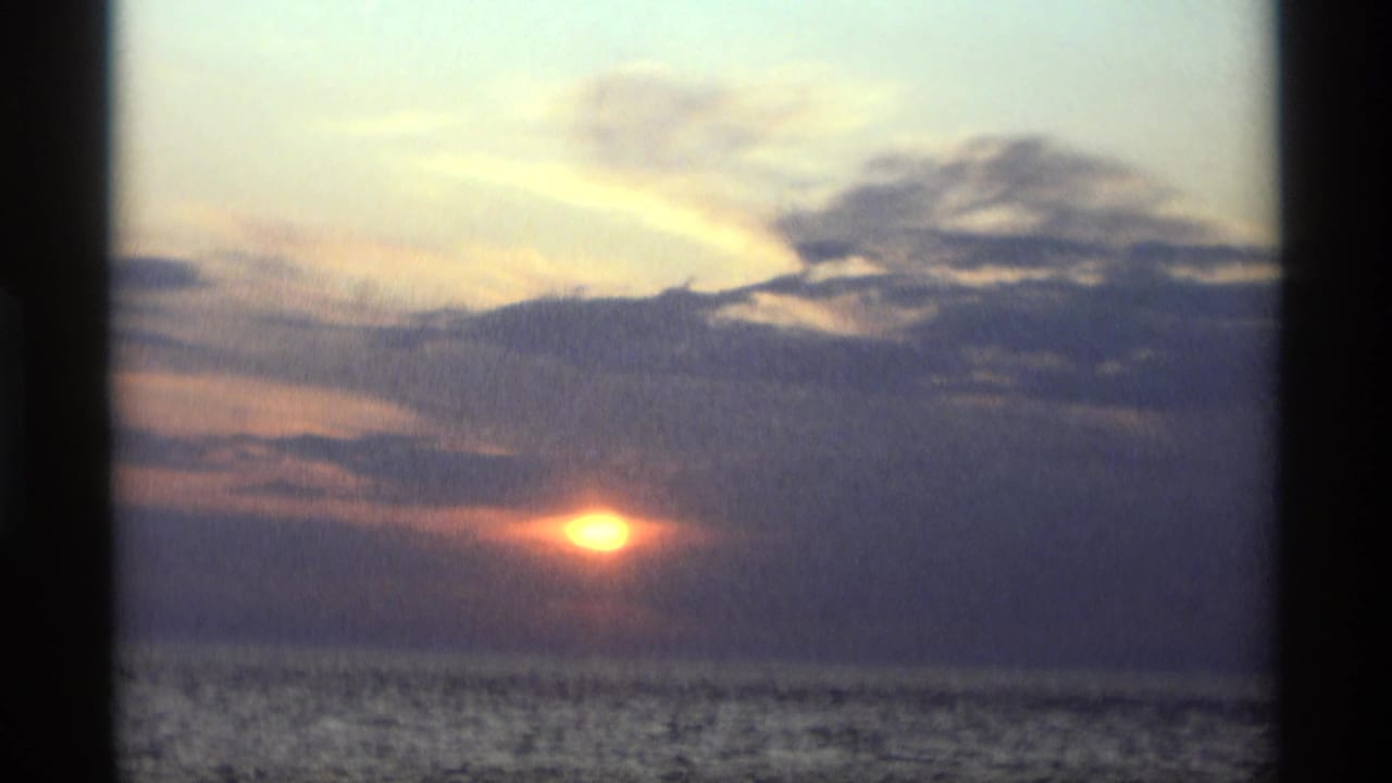 Gold sun near the horizon behind grey clouds over calm lake water.