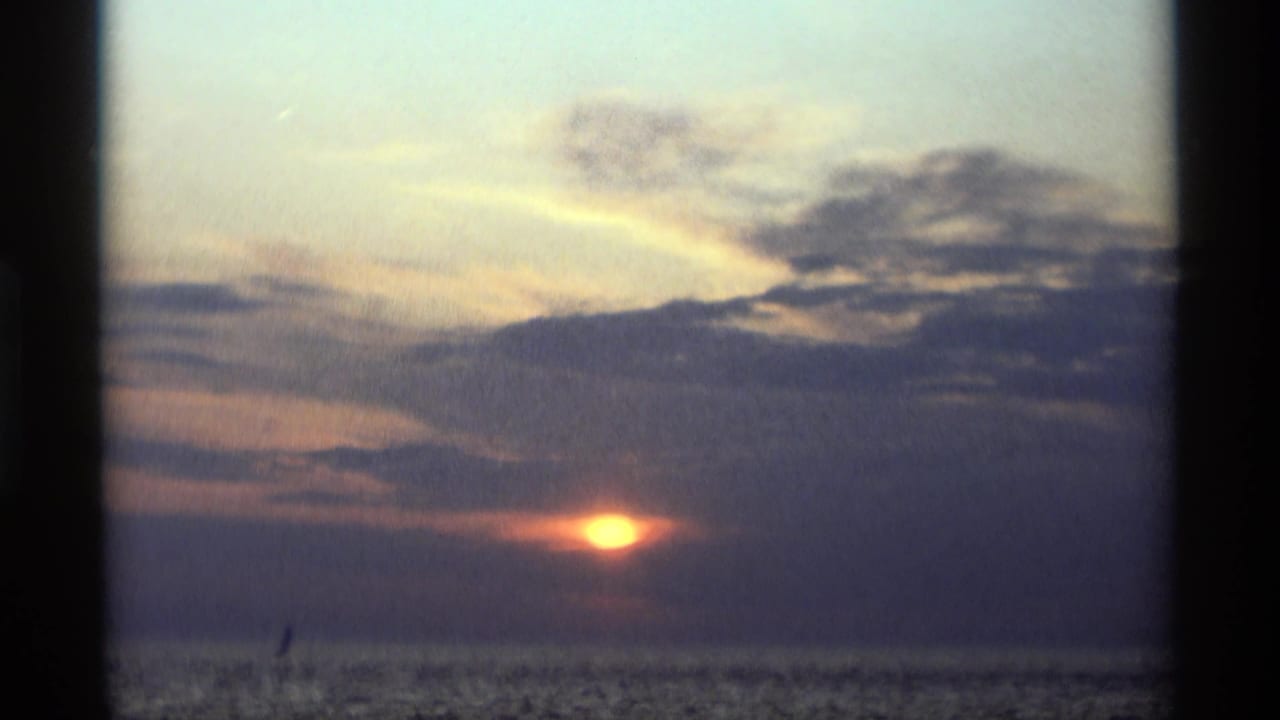 1969 Holland, Michigan lake sunset with the sun partly veiled by clouds and a small sailboat silhouette on the water.