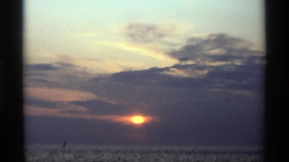 1969 Holland, Michigan lake sunset with the sun partly veiled by clouds and a small sailboat silhouette on the water.