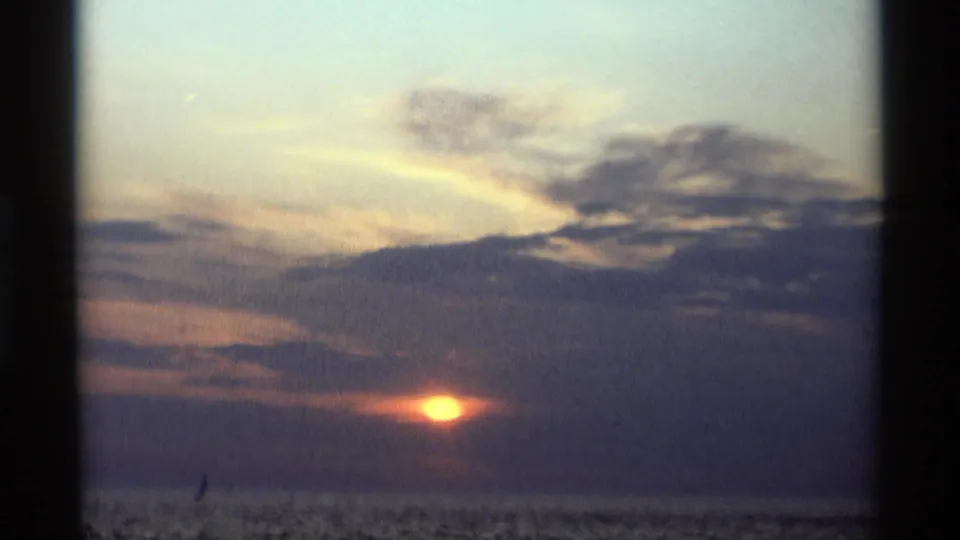 A late-1960s 8mm view of the sun dipping into a grey cloud layer over calm water in Holland, Michigan.