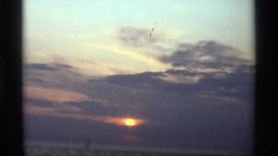 1969 8mm Holland, Michigan sunset frame with a small gold sun below heavy clouds over still lake water.