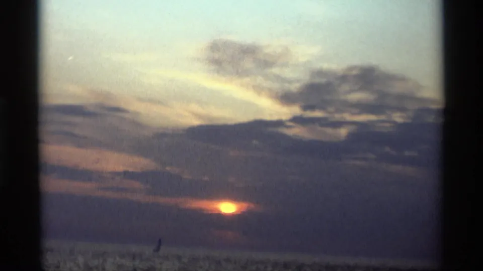 1969 Holland, Michigan 8mm frame showing the sun low behind clouds over water, with a tiny sail silhouette near the horizon.