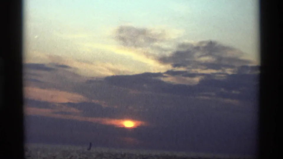 1969 Holland, Michigan 8mm frame of a sunset over open water, framed by dark film edges and a small distant sail silhouette.