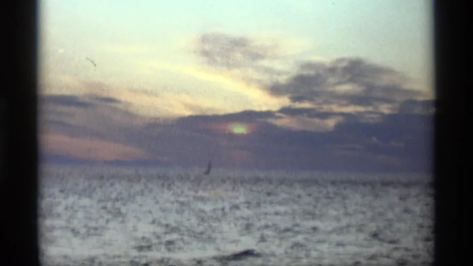 1969 Holland, Michigan 8mm frame with the sun diffused by clouds above rippled water and a small sail silhouette offshore.