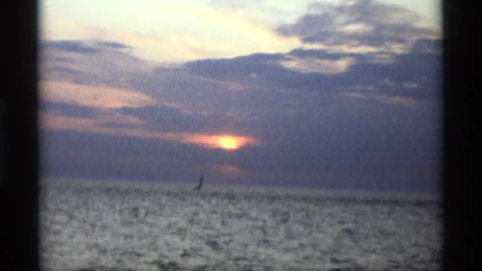 1969 Holland, Michigan 8mm frame showing an orange sun behind cloud cover over open water, with a small distant sail silhouette.