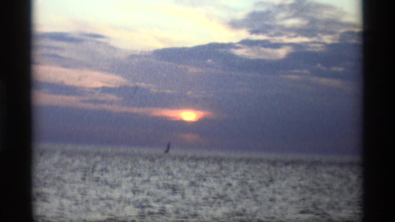 1969 Holland, Michigan home-movie frame showing the sun low over open water, cloud bands, and a distant sailboat silhouette.