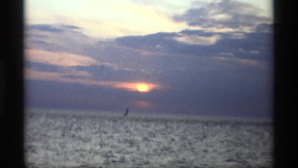 1969 Holland, Michigan home-movie frame showing the sun low over open water, cloud bands, and a distant sailboat silhouette.