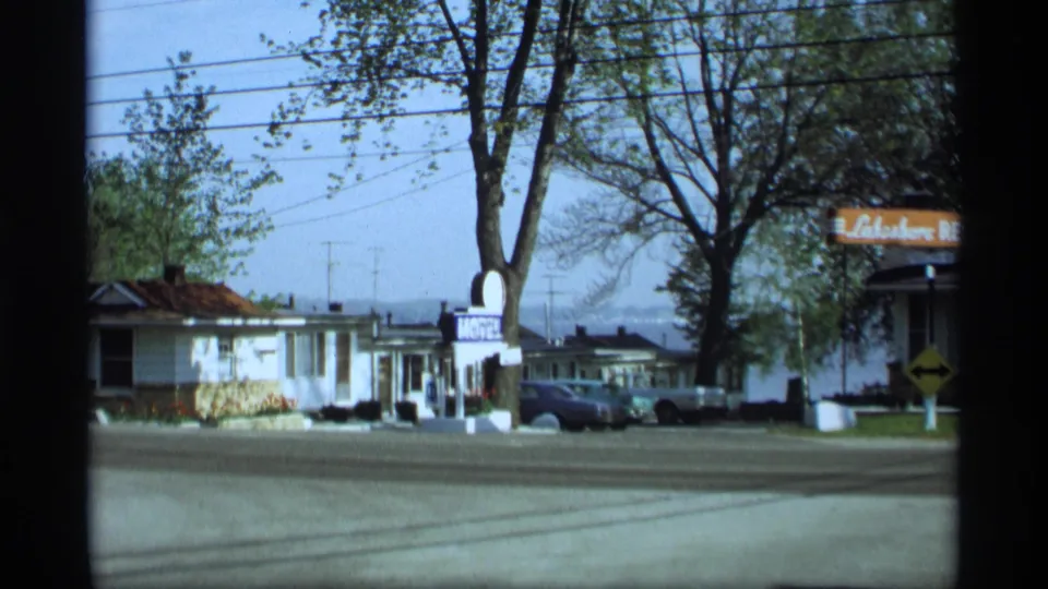 1969 Holland, Michigan roadside frame with a motel sign, parked cars, low buildings, tall trees, and overhead wires.