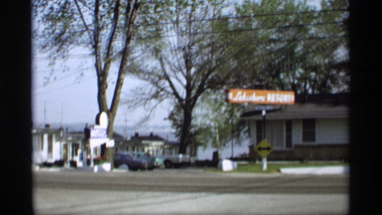 Roadside view of a resort sign, low buildings, parked cars, trees, and street.