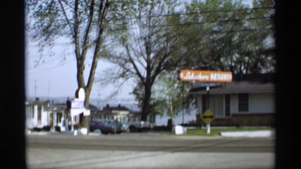 1969 Holland, Michigan 8mm frame showing a roadside resort sign near low buildings, parked cars, trees, and an open street.