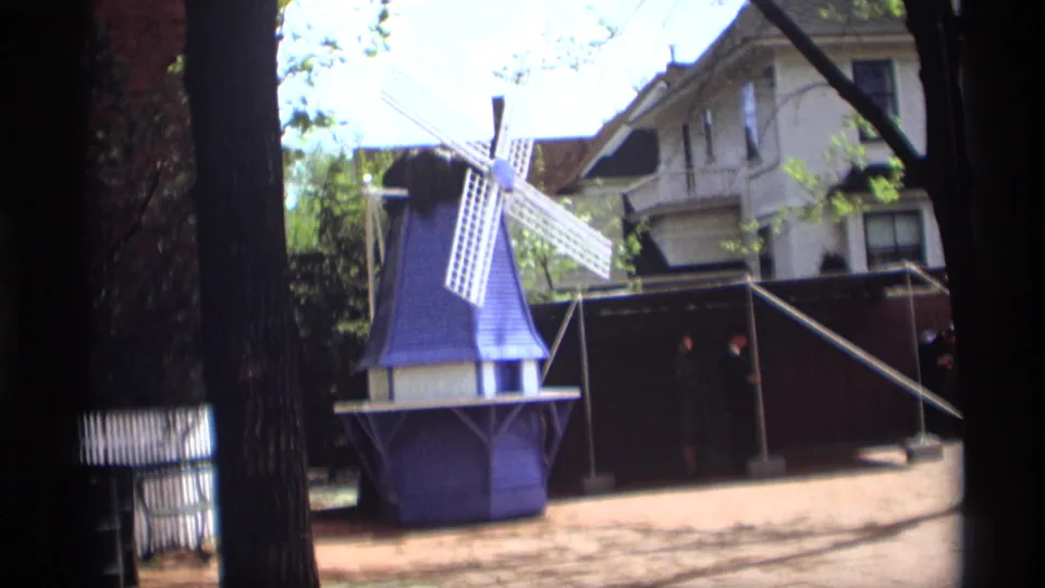 A 1969 home-movie view of a blue Dutch-style windmill display in Holland, Michigan, with trees and residential buildings behind it.