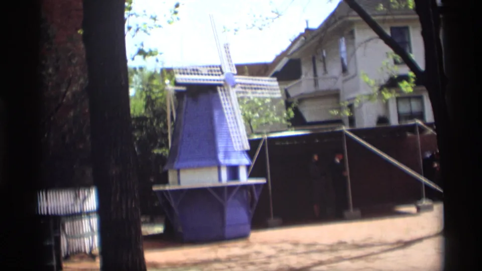 Closer 8mm view of the Holland, Michigan windmill display, with white blades, trees, fencing, and a house visible in the background.
