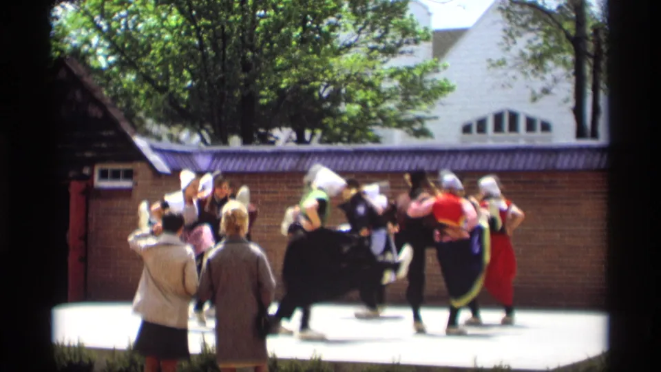 1969 Holland, Michigan 8mm frame showing adults in Dutch-style folk costumes dancing outdoors as spectators watch near a brick wall.