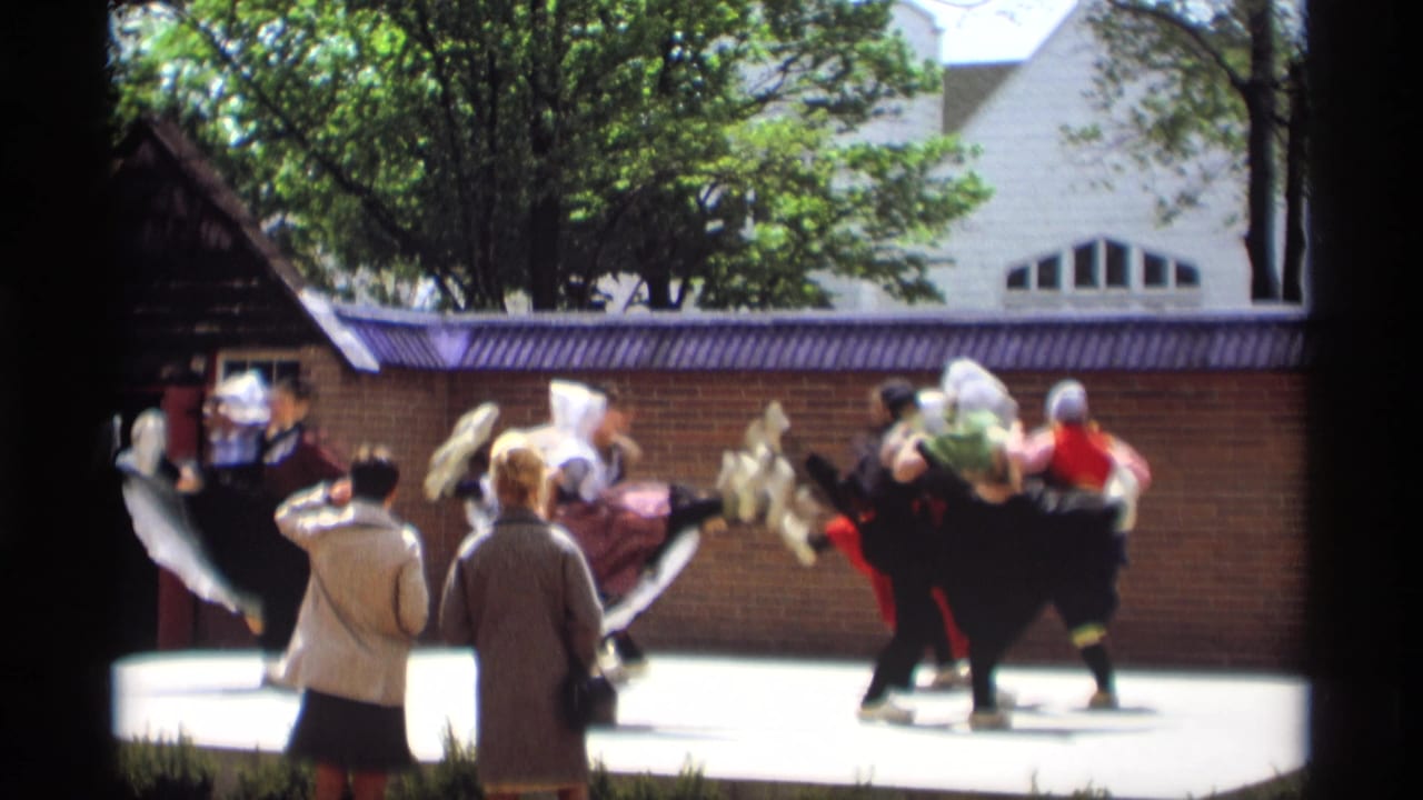 Costumed dancers perform outdoors while two spectators watch from the foreground.