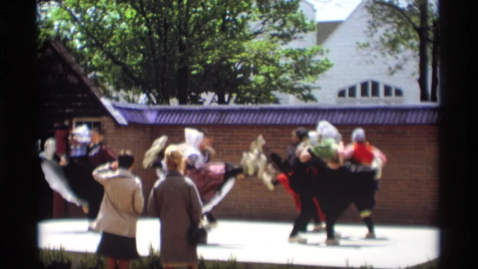 1969 Holland, Michigan home-movie frame of Dutch-style folk dancers moving across a sunlit outdoor stage as spectators watch.