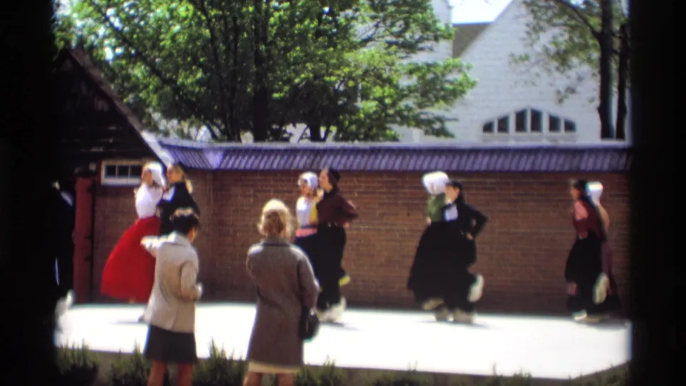 A 1969 outdoor folk dance moment in Holland, Michigan, with bonneted dancers, a red skirt, and foreground spectators in 1960s clothing.