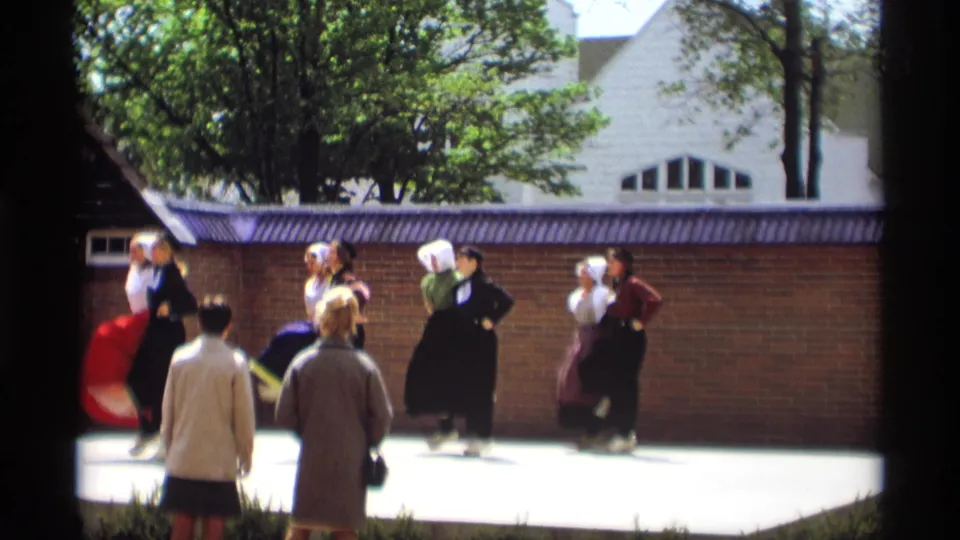 1969 Holland, Michigan home-movie frame of paired Dutch folk dancers on an outdoor stage with spectators in front.