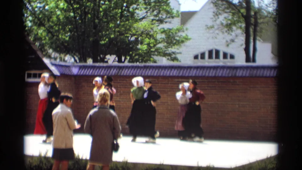 1969 Holland, Michigan frame of Dutch-style dancers in pairs, seen past foreground spectators at an outdoor performance.
