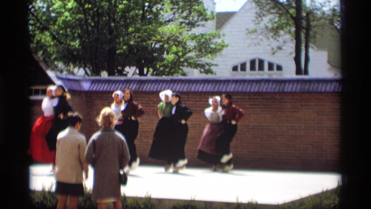 Women in Dutch-style folk costumes continue a synchronized outdoor dance in Holland, Michigan, with onlookers close to the performance area.