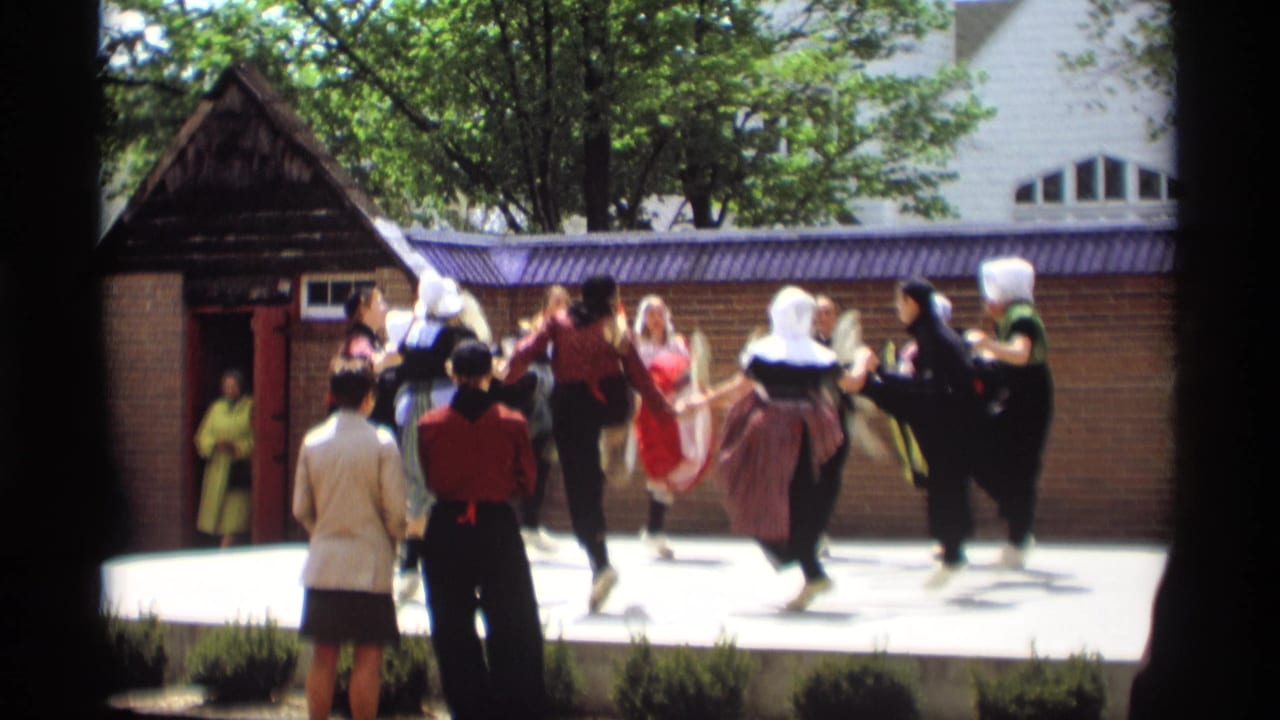 Costumed dancers hold hands on an outdoor stage as two spectators watch from the foreground.