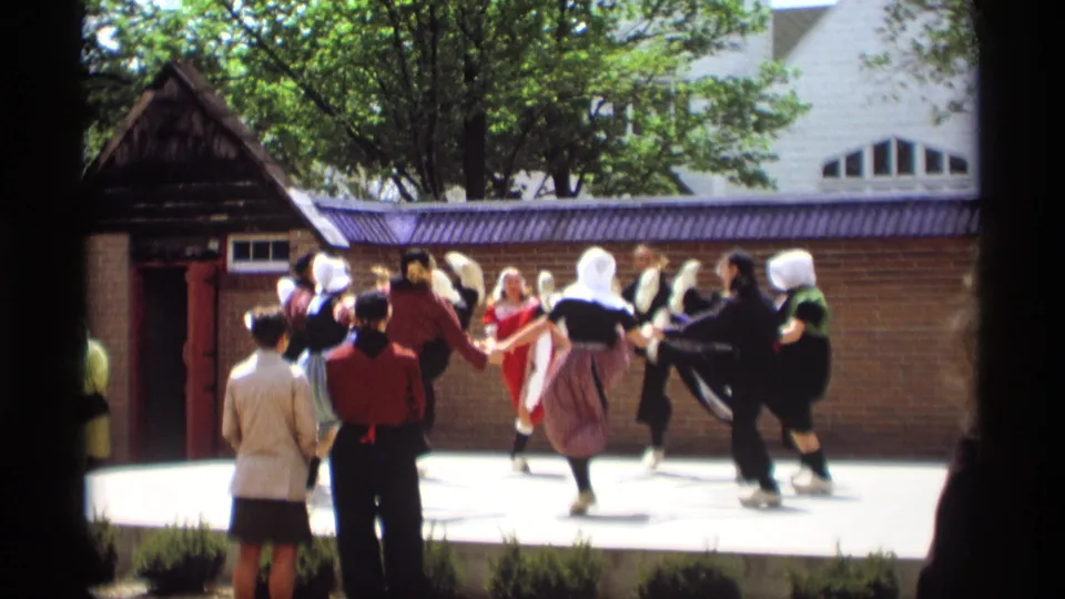 Dutch-style folk dancers in Holland, Michigan continue a circle dance with raised kicks on a bright outdoor stage in 1969.