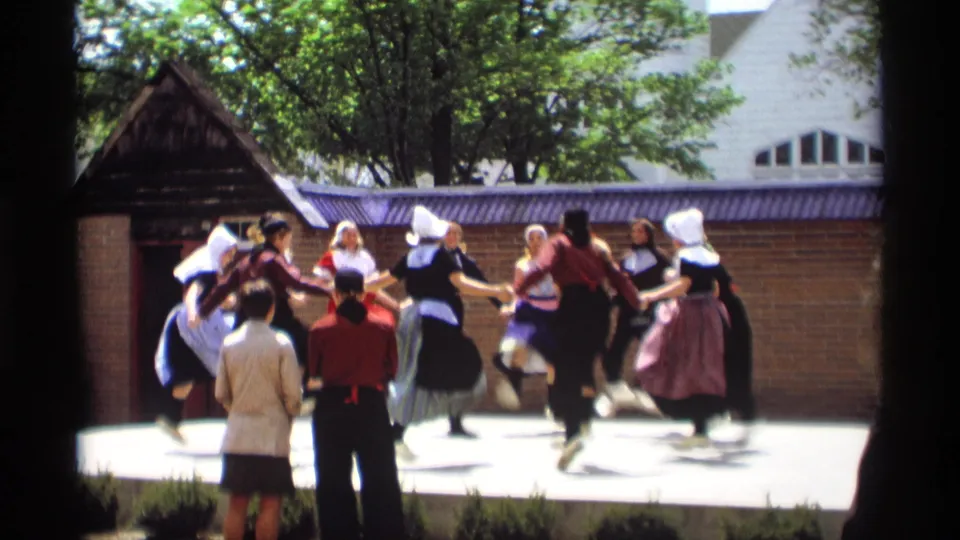 The home-movie view shows the dancers mid-routine, with foreground spectators and a brick wall behind the Holland, Michigan stage.