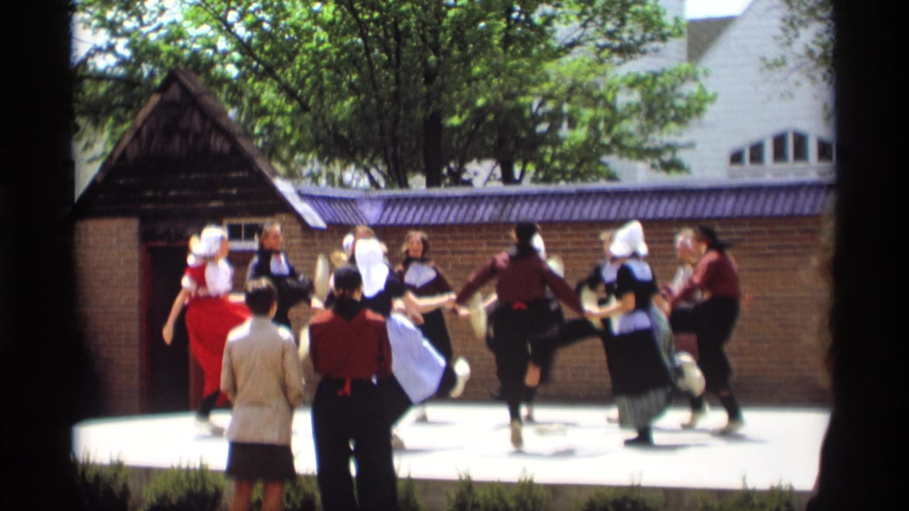 Women in Dutch-style costumes dance in a circle on an outdoor stage as spectators watch.
