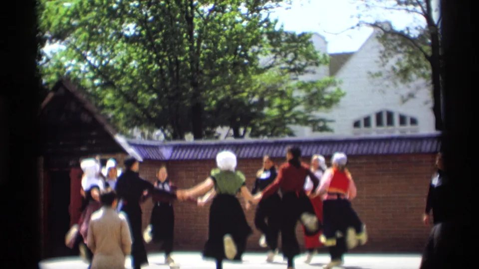 Women in traditional Dutch-style dress continue a circle dance in Holland, Michigan, with spectators partly blocking the lower frame.