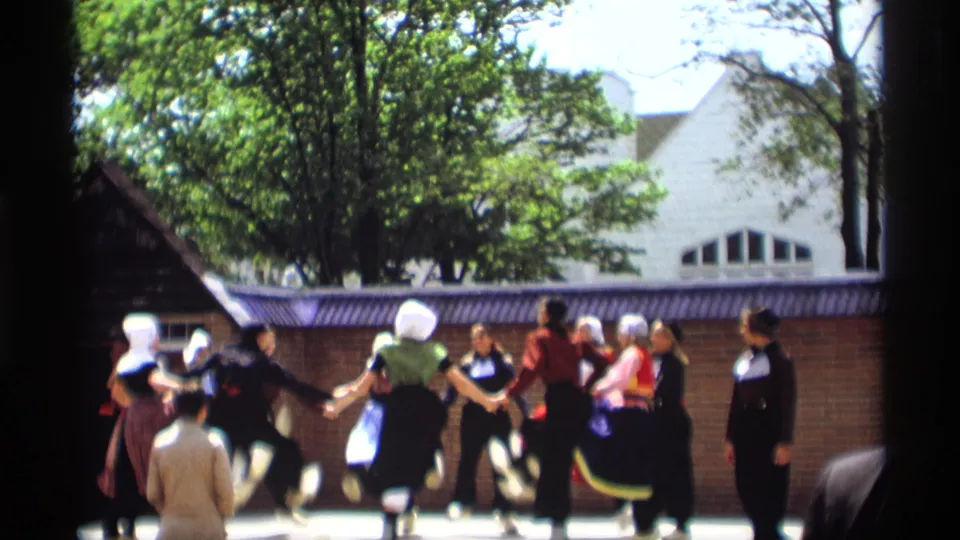 Home-movie view of Dutch-costumed women dancing outdoors in 1969 Holland, Michigan, with trees and buildings behind the stage.
