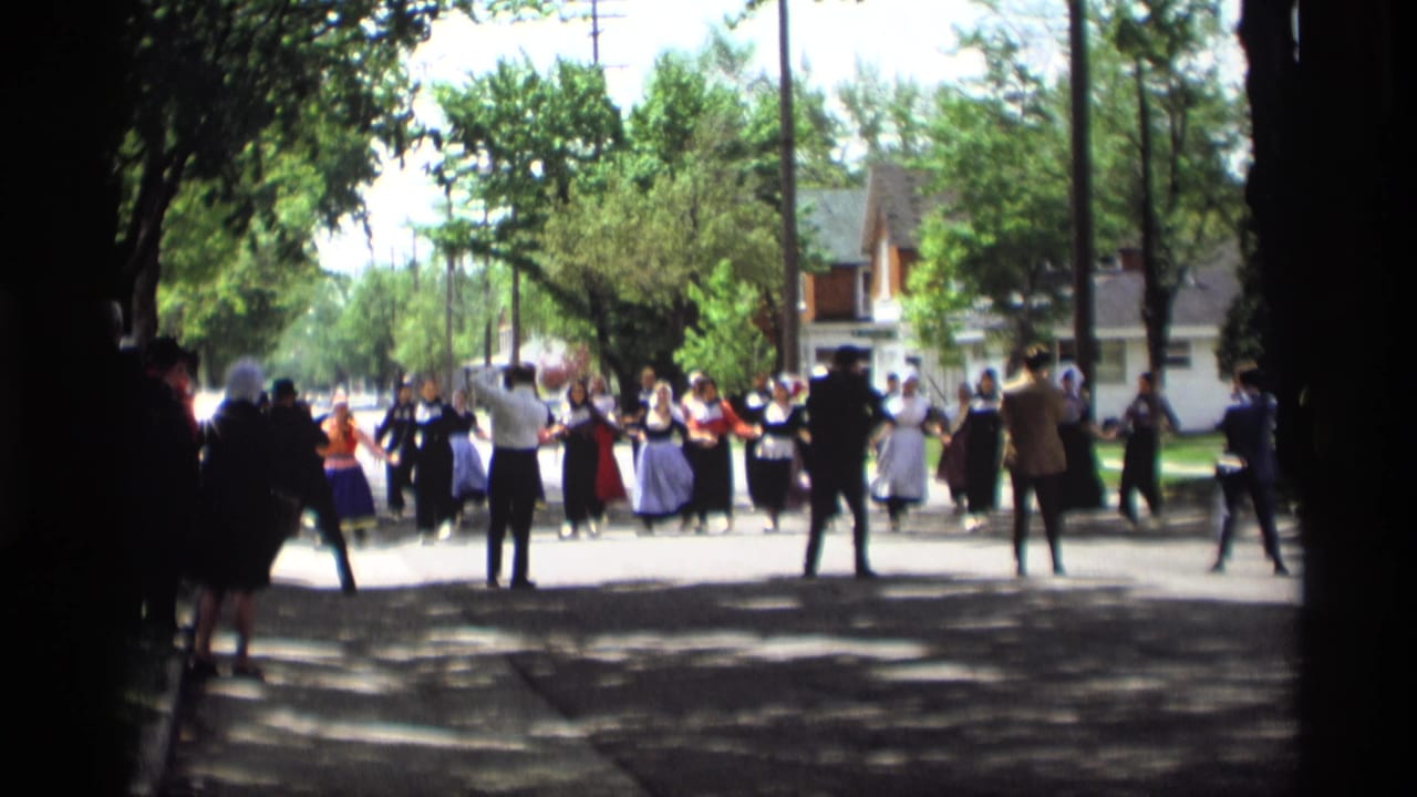 Costumed dancers and suited men perform in a shaded residential street.