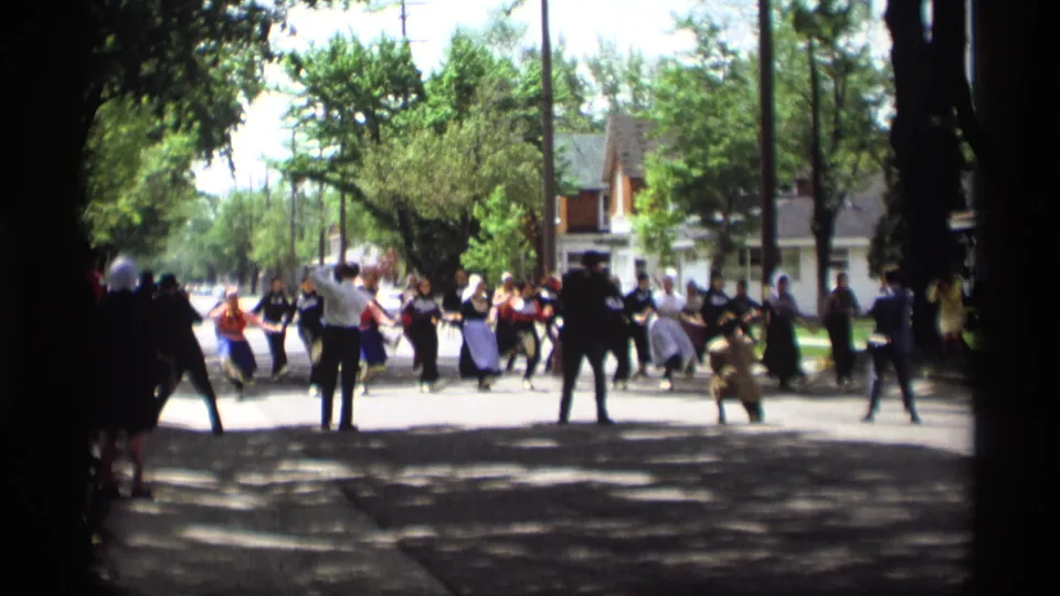 Costumed dancers and men in dark suits continue a coordinated folk dance on a Holland, Michigan residential street in 1969.