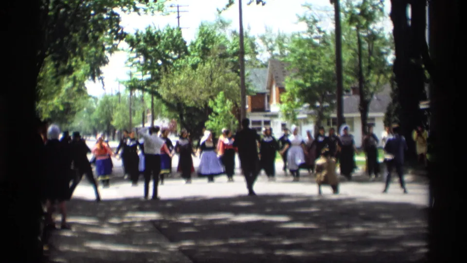 Home-movie view of a 1969 Holland, Michigan street folk dance, with participants moving through shade between foreground tree trunks.