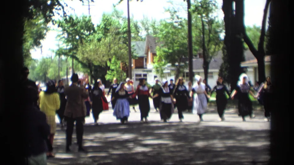 Costumed women and suited men advance in a folk-dance formation on a Holland, Michigan street in this 1969 8mm frame.