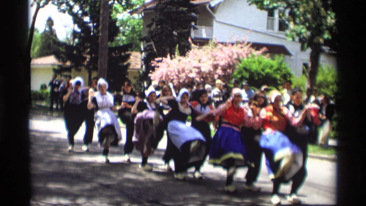 In Holland, Michigan, women in bonnets, aprons, and colorful skirts dance together on a residential street in 1969 home-movie footage.