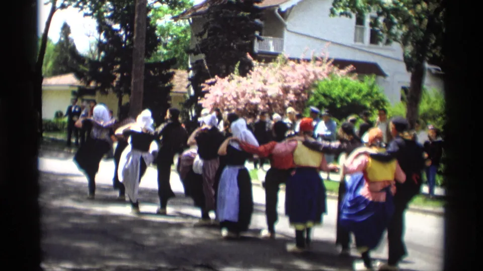 Costumed folk dancers in Holland, Michigan continue down the street, with white bonnets, dark vests, aprons, and spectators visible nearby.