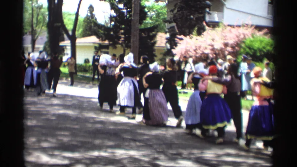 The folk-dance group moves away down a shaded Holland, Michigan street, showing colorful skirts, aprons, bonnets, trees, and nearby spectators.