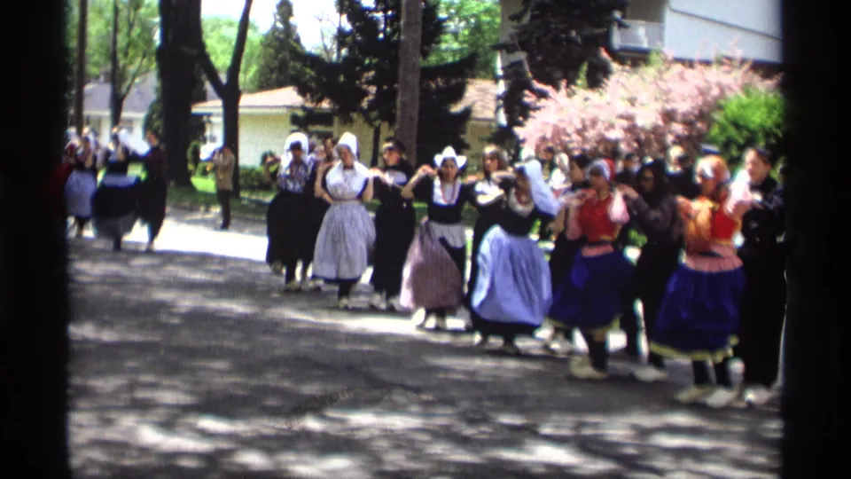 Women in Dutch-style parade costumes walk in formation in Holland, Michigan, with spectators and flowering shrubs visible behind them.
