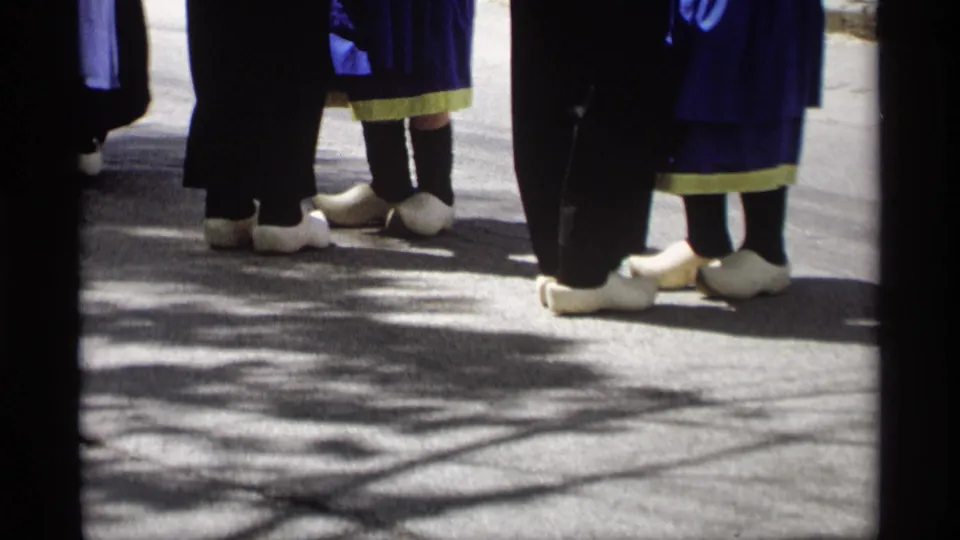 The frame shows the lower half of Dutch-style parade costumes, with white clogs and long skirts grouped on the shaded pavement.